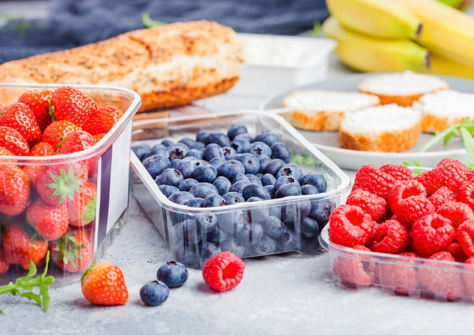 Auf einem Tisch stehen durchsichtige Plastikbehälter, die mit Erdbeeren, Blaubeeren und Himbeeren gefüllt sind. Im Hintergrund befinden sich Brotscheiben, Bananen und mit Frischkäse bestrichene Brotstücke.