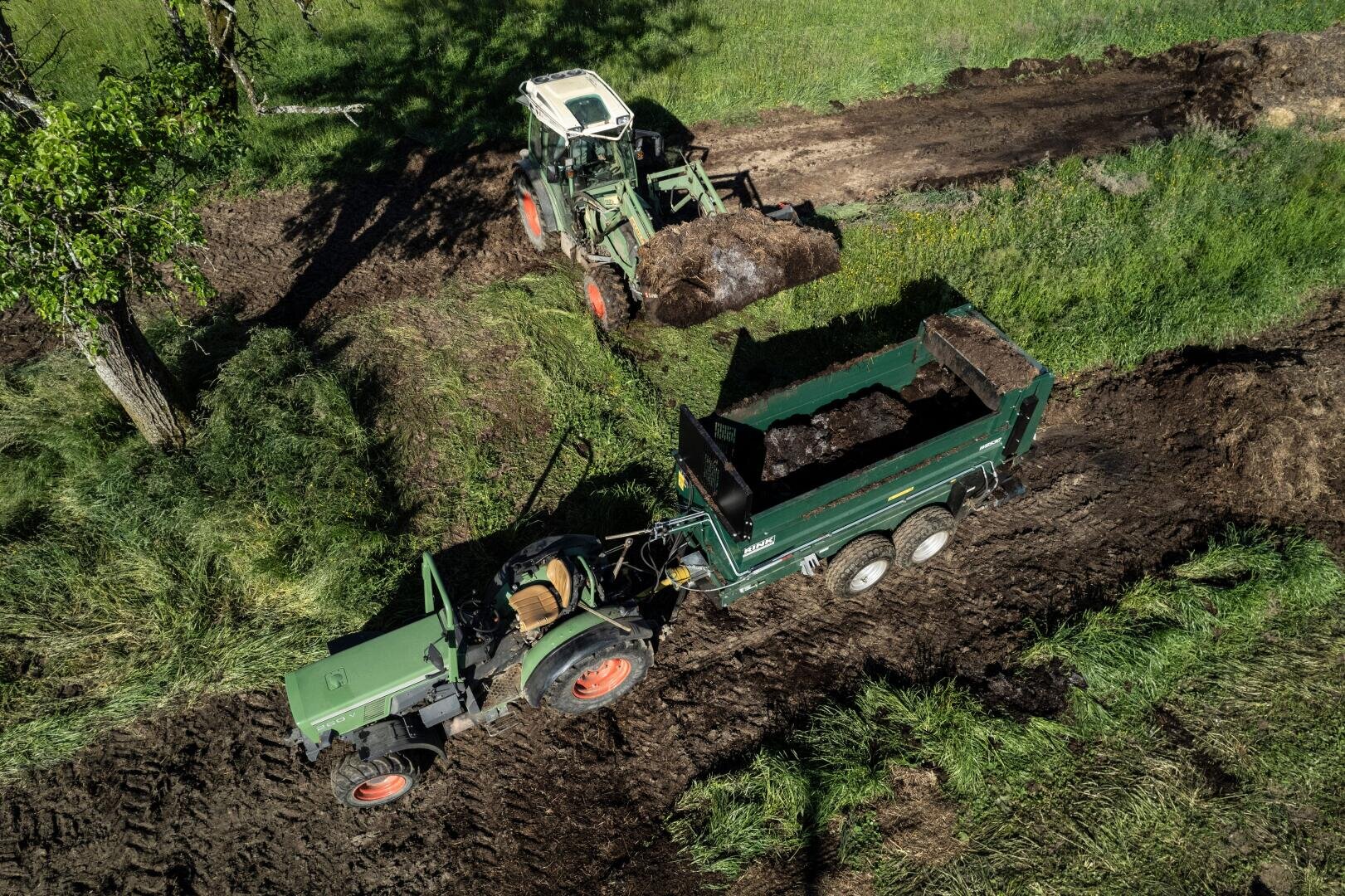 Aerial view of two tractors working on a muddy dirt road in a grassy field. One tractor is loading soil or compost into a green trailer attached to the other tractor.