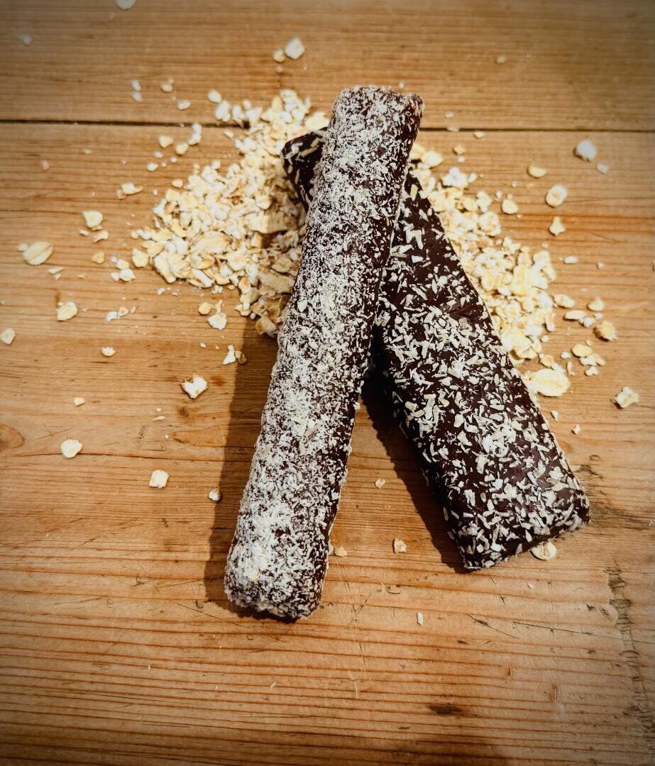 Zwei mit Schokolade überzogene und mit Kokosraspeln bestreute Snackriegel liegen auf einer Holzplatte, dahinter ein Haufen Haferflocken.