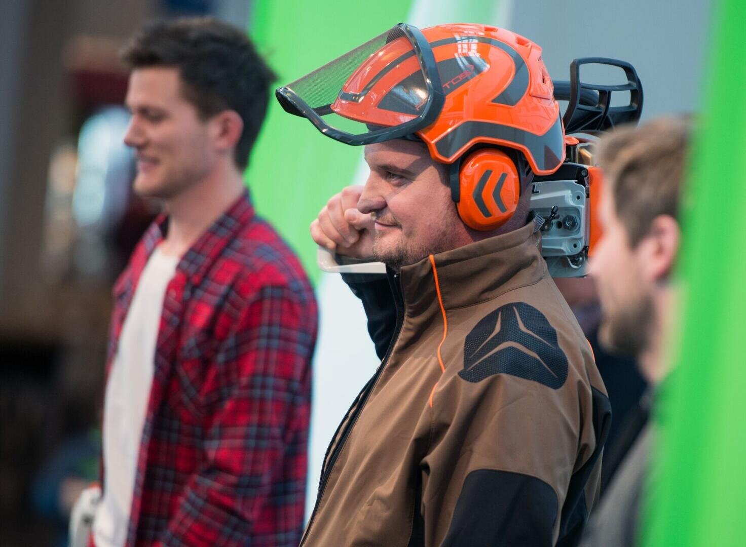 Drei Männer stehen in einem Haus; der Mann in der Mitte trägt eine braune Jacke, einen orangefarbenen Helm mit Gesichtsschutz und stützt eine Kettensäge auf seine Schulter. Die beiden anderen Männer sind im Vorder- und Hintergrund unscharf.