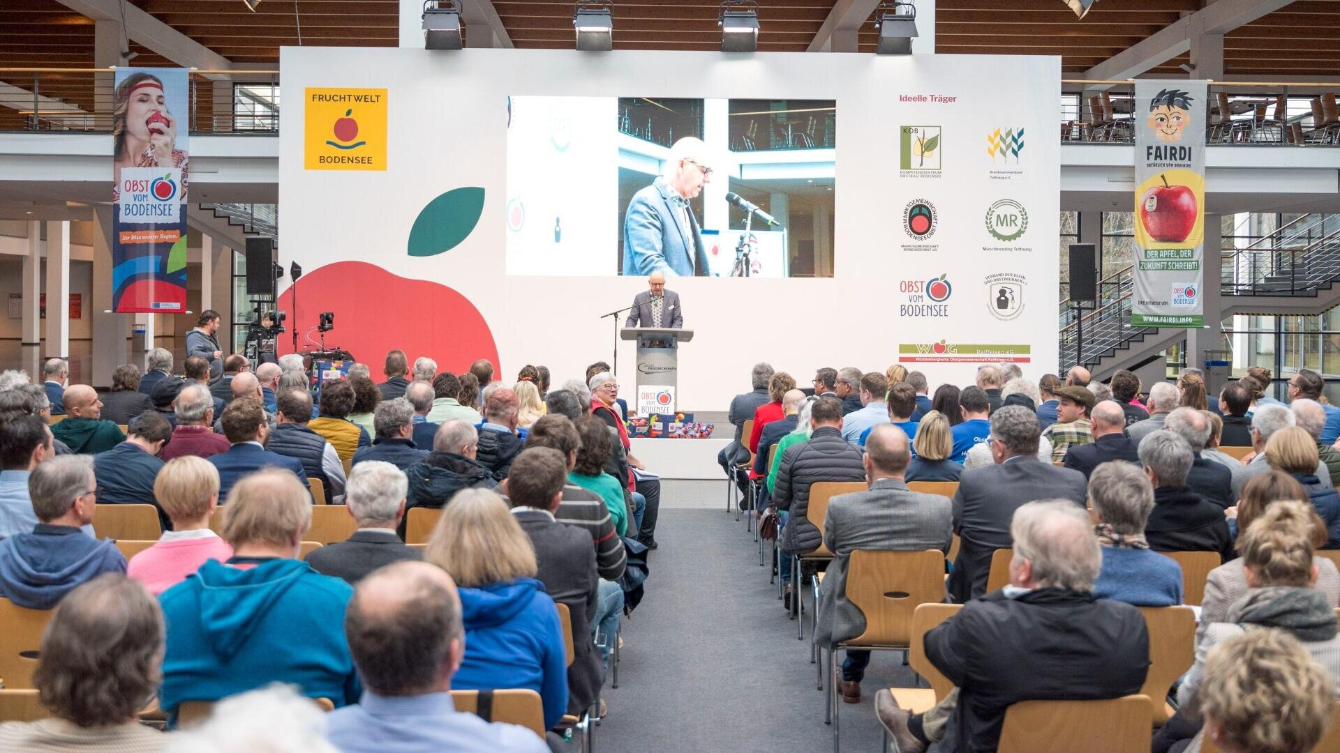 Ein großes Publikum sitzt vor einer Bühne, auf der ein Mann auf einem Podium spricht. Hinter ihm befindet sich eine Leinwand mit seinem Bild und Bannern mit Logos von Obst. Die Veranstaltung findet in einer hellen, modernen Halle statt.