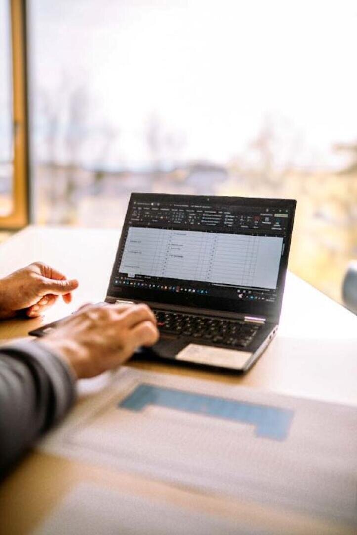 Eine Person arbeitet an einem Laptop mit einer Tabellenkalkulationsanwendung an einem hellen Schreibtisch in der Nähe eines Fensters, wobei im Vordergrund unscharfe Dokumente zu sehen sind.