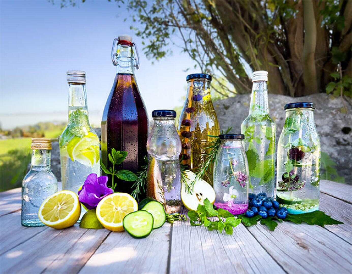 Various glass bottles filled with colorful water, herbs, fruit and flowers are placed on a wooden table outside. Sliced lemons, cucumbers, blueberries and leaves are scattered around the bottles.