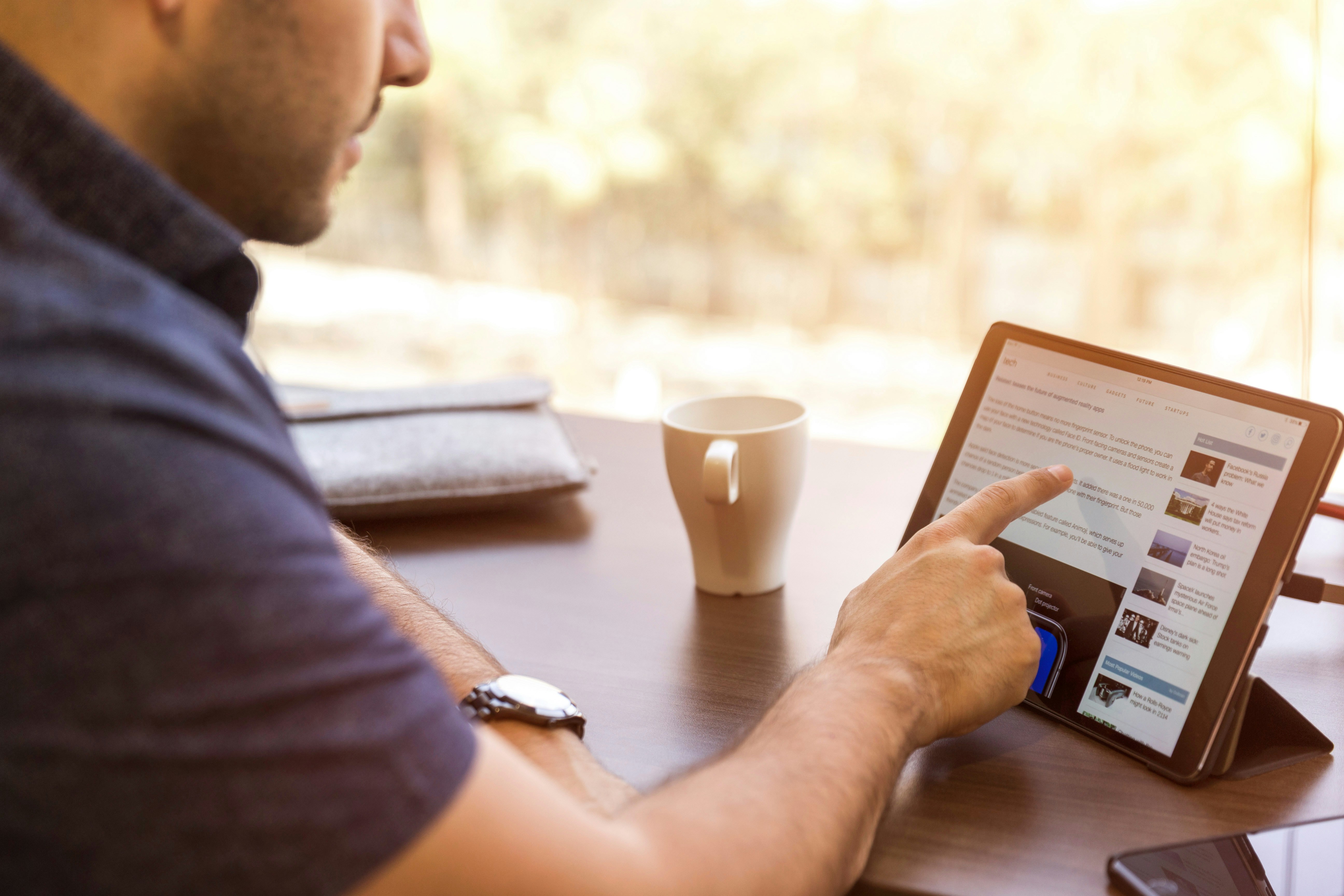 A person sits at a desk using a tablet, browsing news or articles. A coffee mug and closed laptop are on the table, with sunlight streaming in through a window in the background.