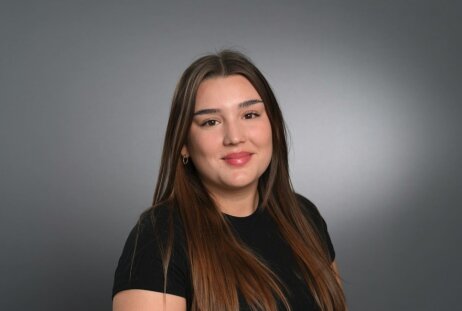 Eine junge Frau mit langen, glatten braunen Haaren und einem schwarzen, kurzärmeligen Hemd lächelt sanft vor einem schlichten grauen Hintergrund.