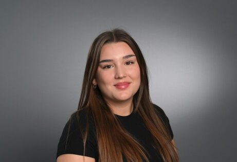 Eine junge Frau mit langen, glatten braunen Haaren und einem schwarzen, kurzärmeligen Hemd lächelt sanft vor einem schlichten grauen Hintergrund.