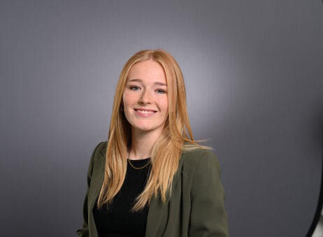 Eine junge Frau mit langen blonden Haaren, die einen dunkelgrünen Blazer über einem schwarzen Oberteil trägt, lächelt vor einem schlichten grauen Hintergrund.