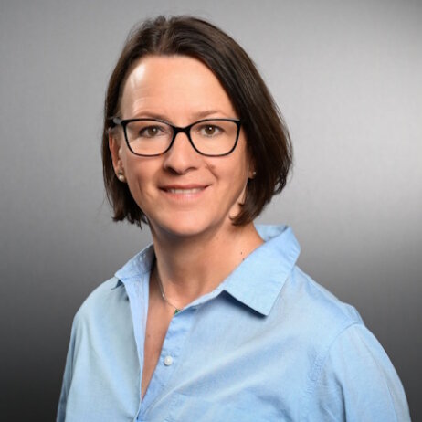 Eine Frau mit kurzen braunen Haaren, die eine Brille und ein hellblaues Hemd mit Kragen trägt, lächelt in die Kamera vor einem schlichten grauen Hintergrund.