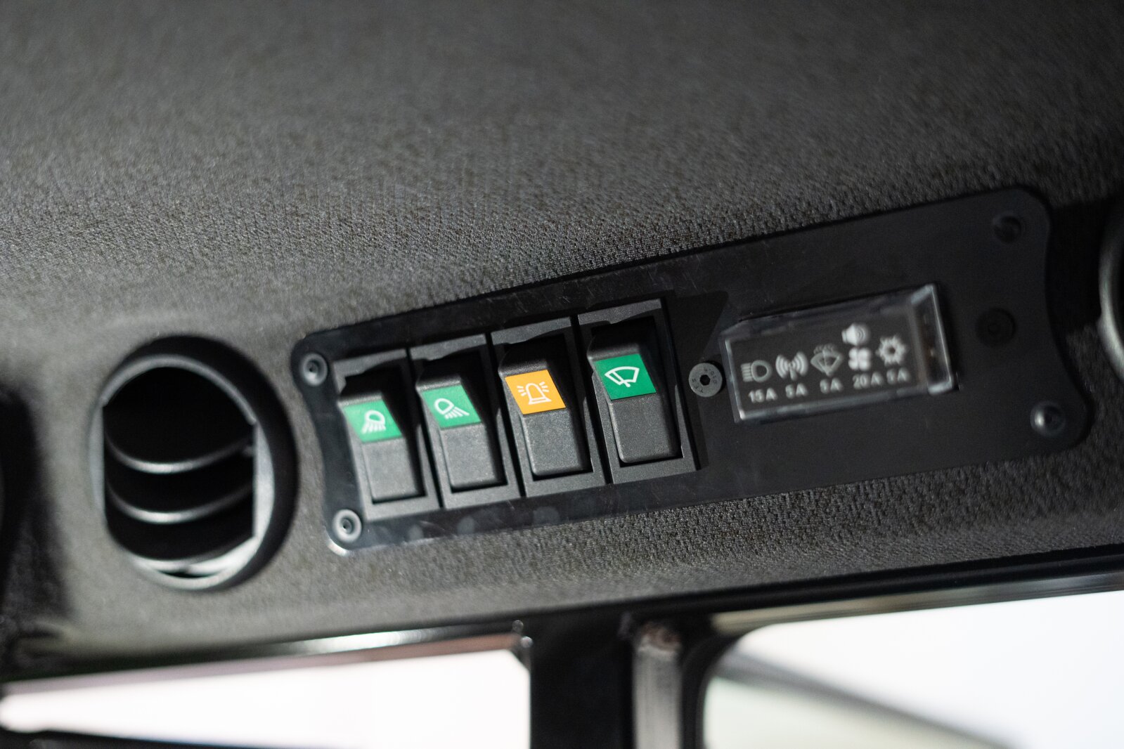 Close-up of a black dashboard with five switches, each marked with green or yellow symbols for different vehicle functions, next to a round ventilation opening.